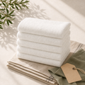 Five white hotel towels styled with sage linen and olive leaves in warm window light